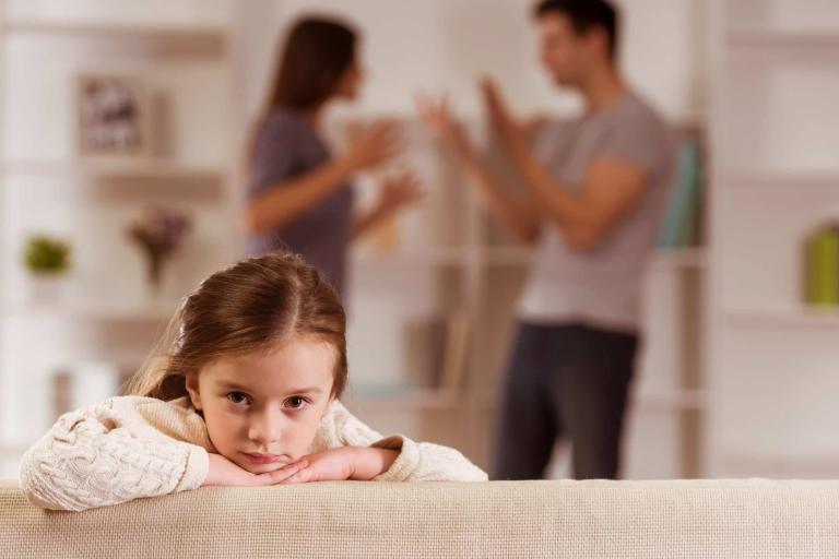 young girl sitting on sofa with parents arguing in background